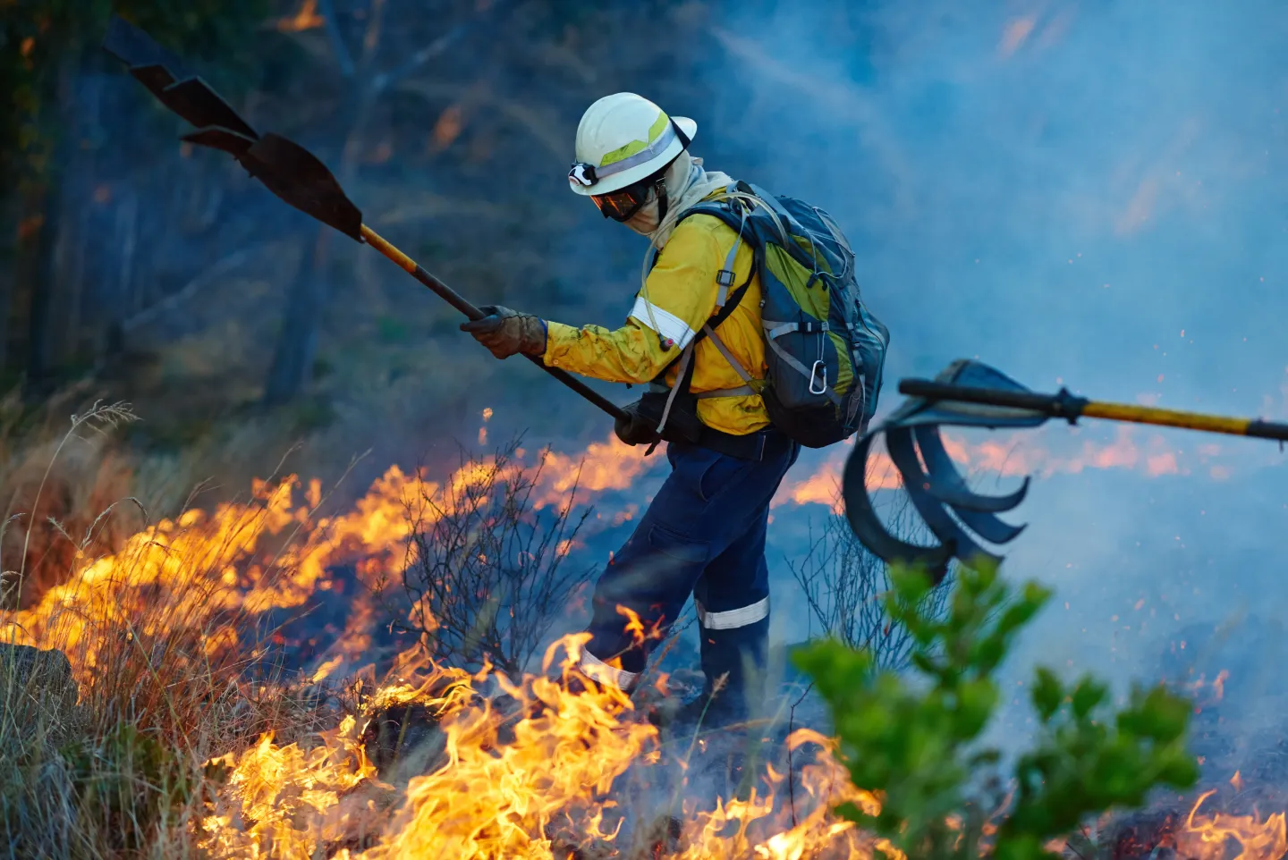 A single firefighter in protective gear working close to an intense wildfire in backcountry terrain.